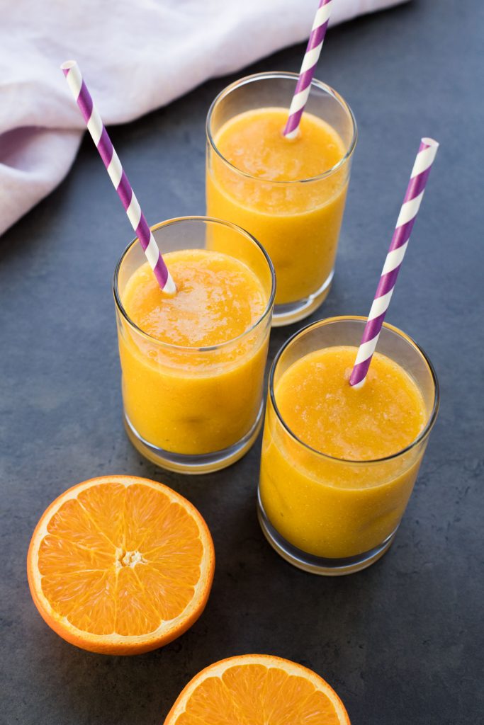 Three glasses of mango orange smoothie with straws and a tablecloth in the background